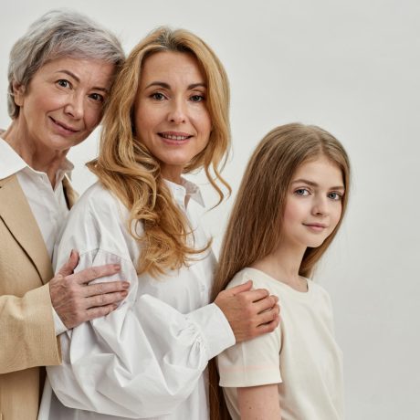 Side view of smiling european family of three female generations hug and look at camera. Age and generation concept. Grandmother, mother and granddaughter. White background. Studio shoot. Copy space
